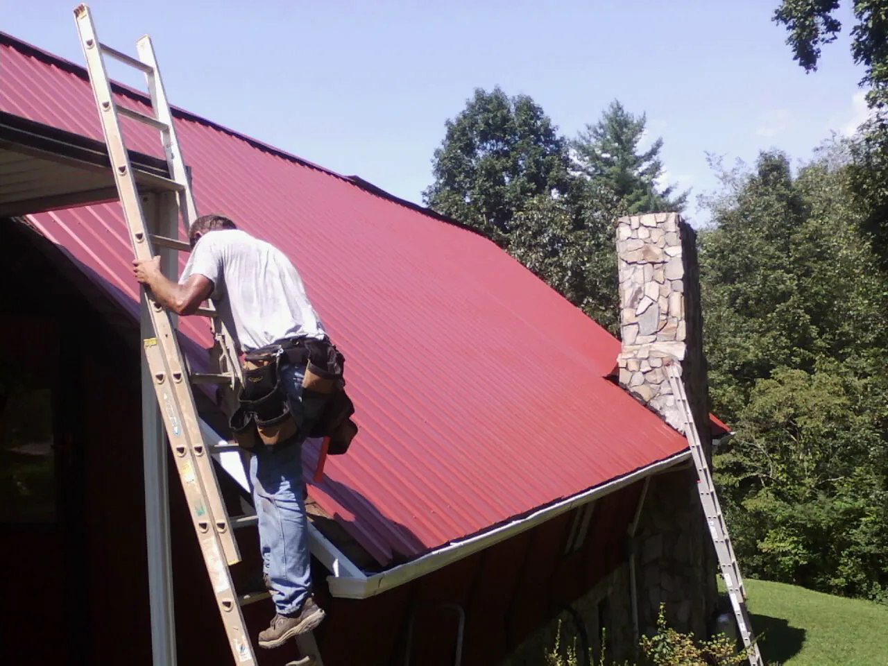 Roofer on a ladder beside a red metal roof and stone chimney — Soffit & Fascia Repair by Roofing Brothers in Lansing